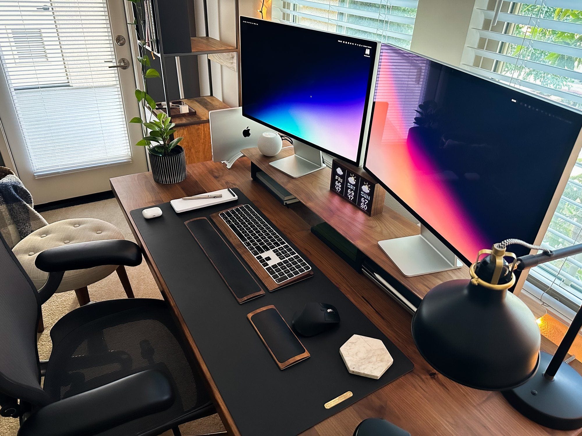 An inviting workspace with a dual monitor setup, featuring an iMac, a MacBook Pro on a desk mat with a wooden accent, a potted plant, a stylish desk lamp, and a comfortable ergonomic chair, all set against a backdrop of a window with white blinds