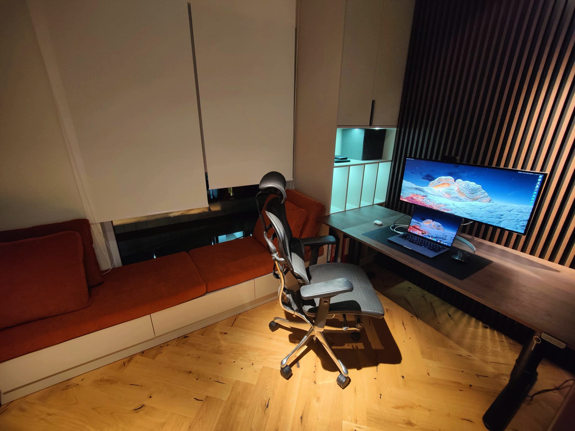 An elegant home office with an adjustable chair positioned before a wooden desk, showcasing a monitor and a laptop, beside a plush orange sofa and white storage units, all under warm ambient lighting