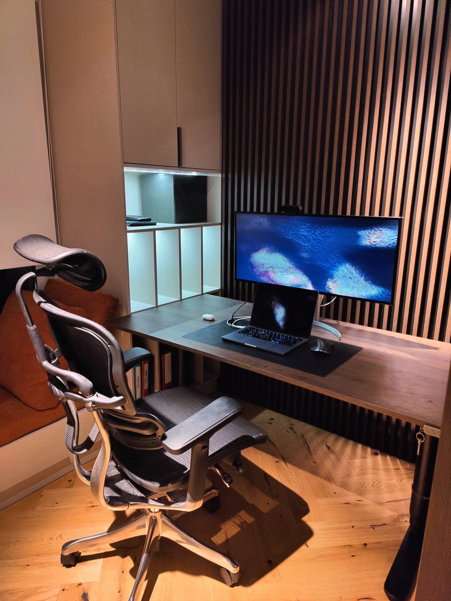 A modern home office with a high-backed ergonomic chair, a wooden desk housing a monitor and laptop, set against a backdrop of warm-toned vertical slatted wall panels and a white shelving unit