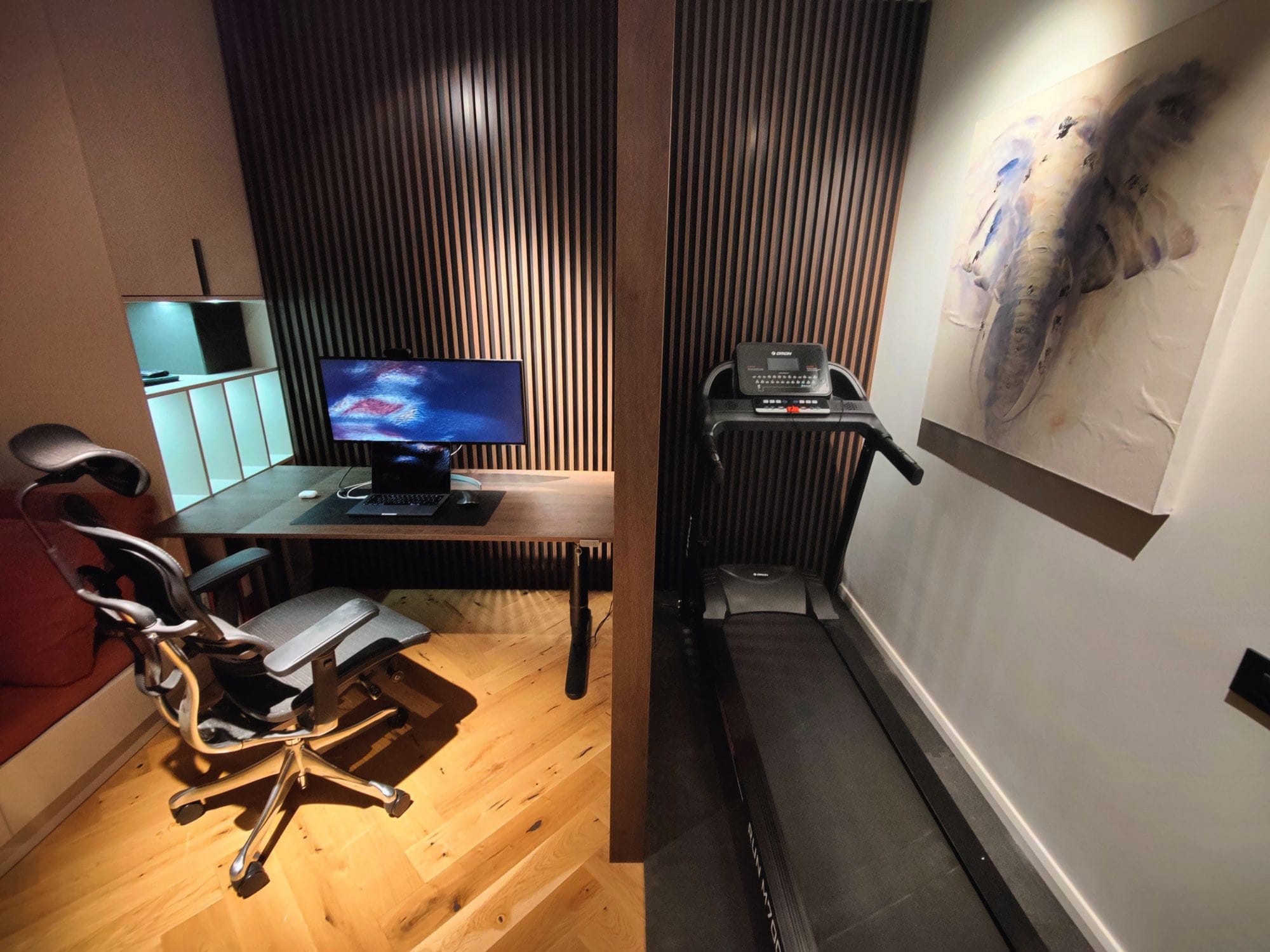 A home office corner with a sleek desk and ergonomic chair, a monitor displaying a cosmic image, adjacent to a treadmill, against a backdrop of vertical wooden slats and an elephant painting