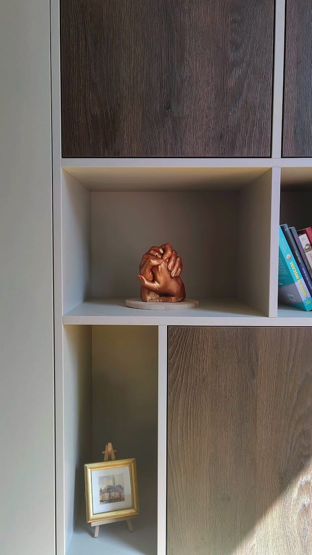 A close-up of a white shelving unit displaying a bronze sculpture of interlocked hands in one compartment, with a small framed picture and a selection of books in the others, all set against a backdrop of natural light