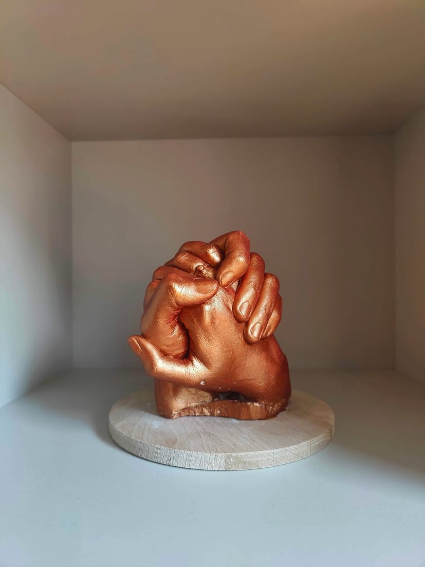 A bronze sculpture of intertwined hands on a circular wooden base, centred within a light beige cubby shelf, highlighted by soft, natural light