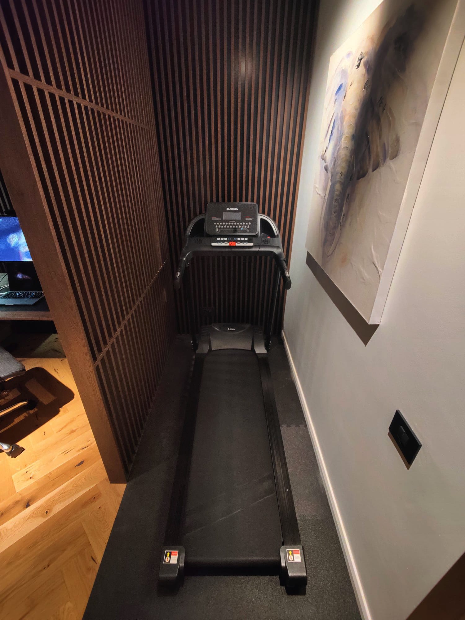 A compact treadmill is positioned against a wall with vertical wooden slats on one side and a large abstract painting on the other, in a room with wooden flooring and soft lighting, suggesting a blend of fitness and art in a home environment