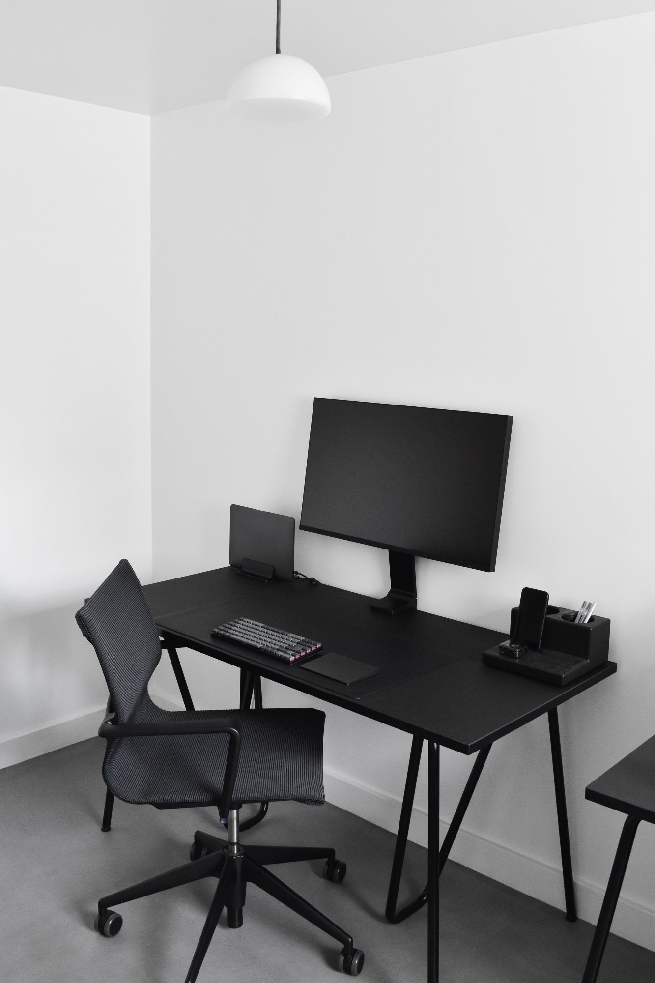 A minimalist black and white home office with a sleek black desk, ergonomic mesh chair, and contemporary desktop accessories under a simple pendant light