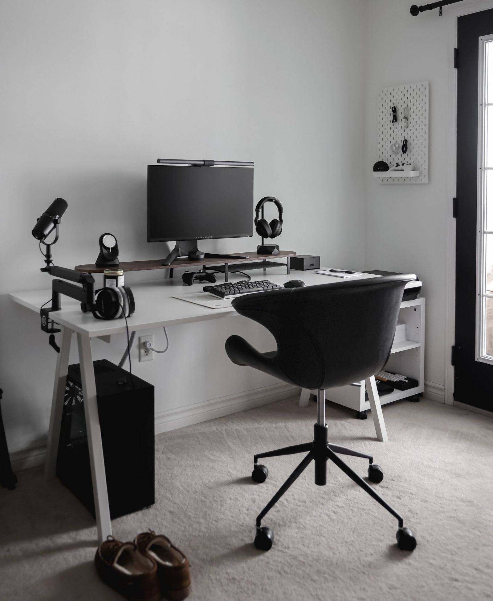 A minimalist home office featuring a white desk, black ergonomic chair, computer monitor, microphone, headphones, and a pegboard with neatly hung accessories against a light grey wall