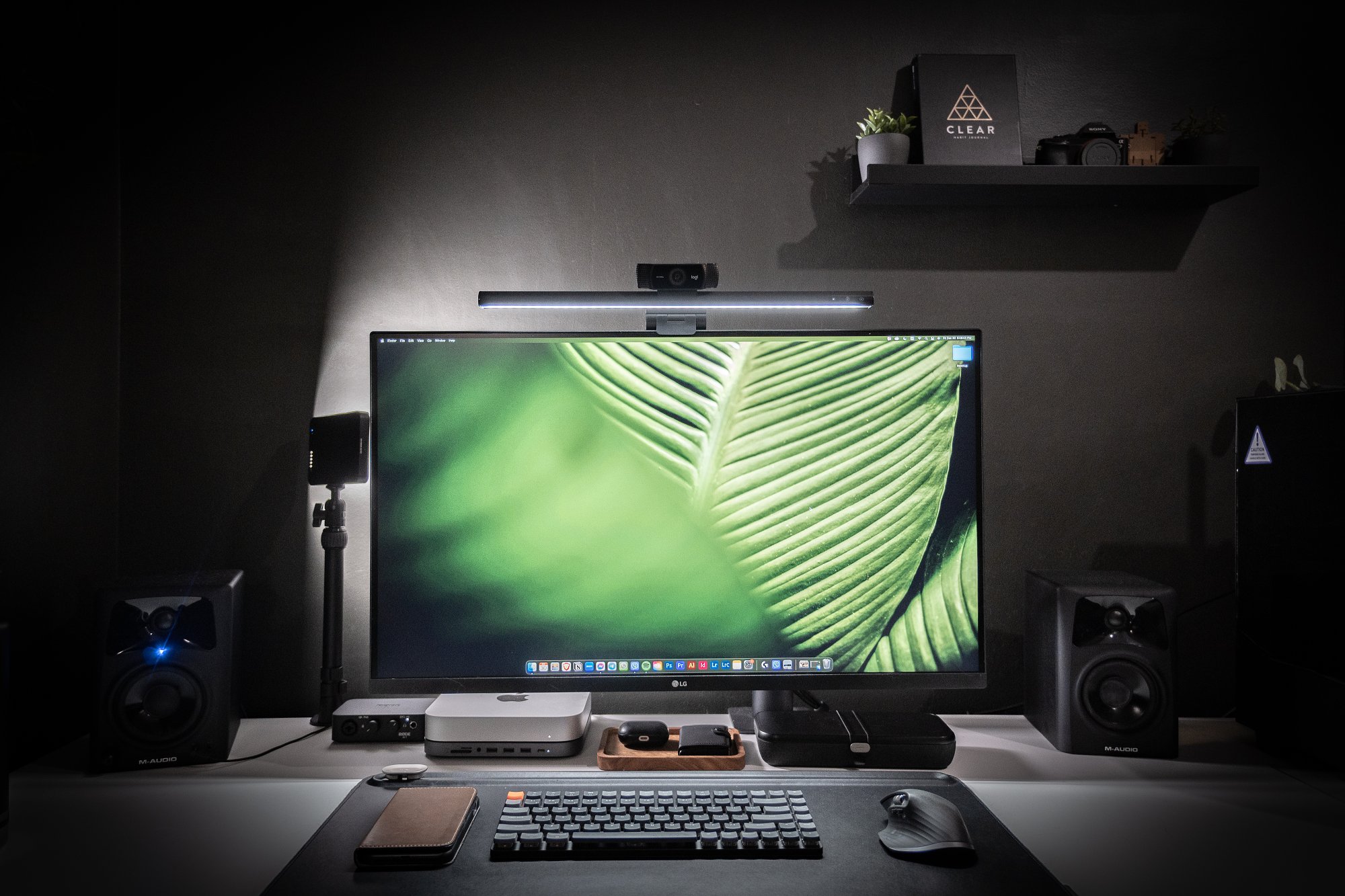 A modern minimalist desk setup with a large monitor, studio speakers, a sound interface, and stylish desktop accessories, all placed on a clean white desk against a dark wall with a decorative shelf above
