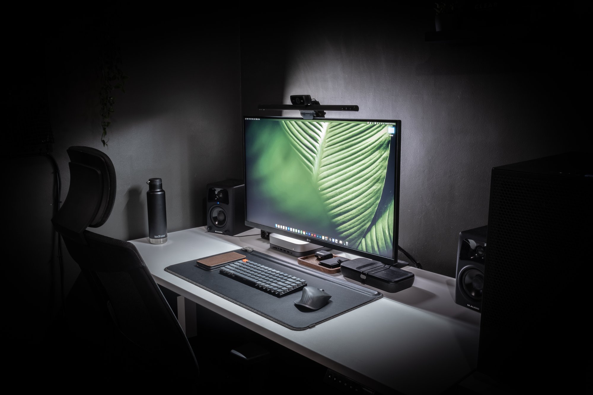 A modern, minimalist home office setup with a large monitor centered on a white desk, flanked by speakers and a plant. The desk has a keyboard, mouse, tablet, and a water bottle, with a comfortable office chair in front. The room has dim lighting focusing on the desk area