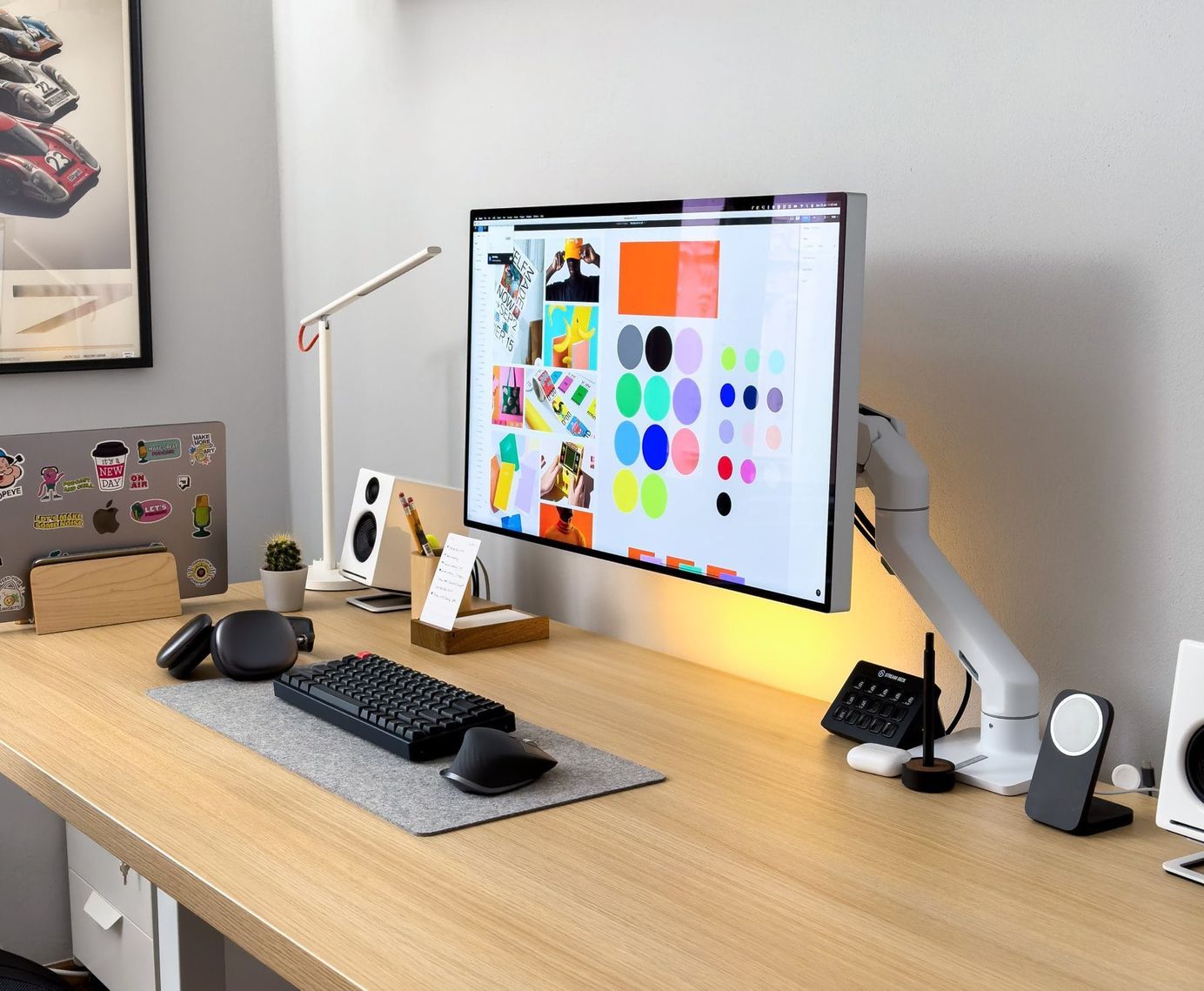 A modern and well-organised minimal desk setup with a large monitor displaying a colourful graphic design project, a sleek white desk lamp, speakers, a black mechanical keyboard and mouse, a notebook, potted plant, and various desk accessories in a bright room