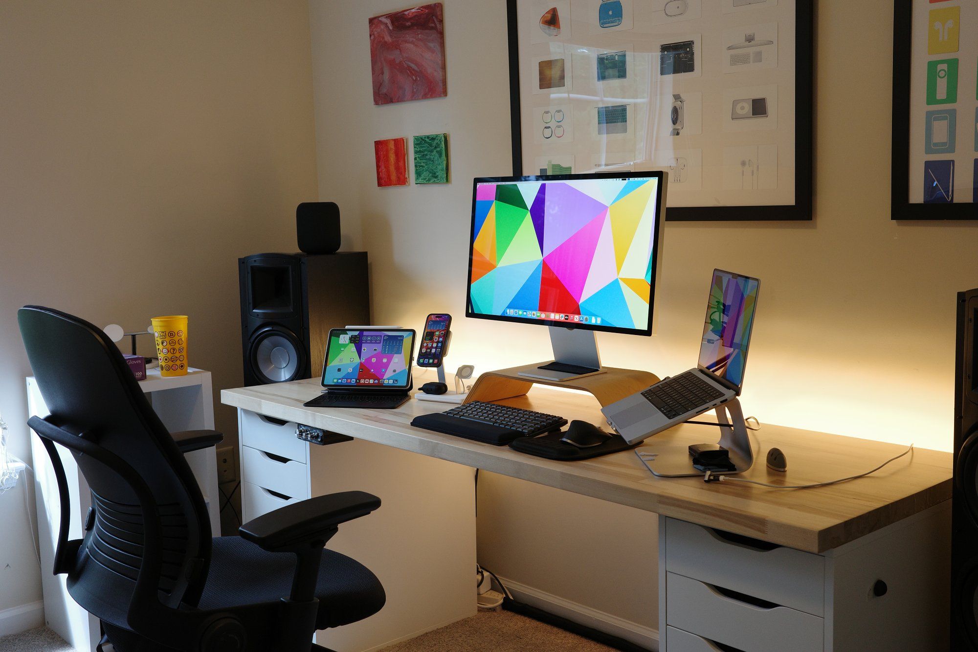 A modern home office setup with a sleek wooden desk featuring an Apple Studio Display with the colourful abstract wallpaper