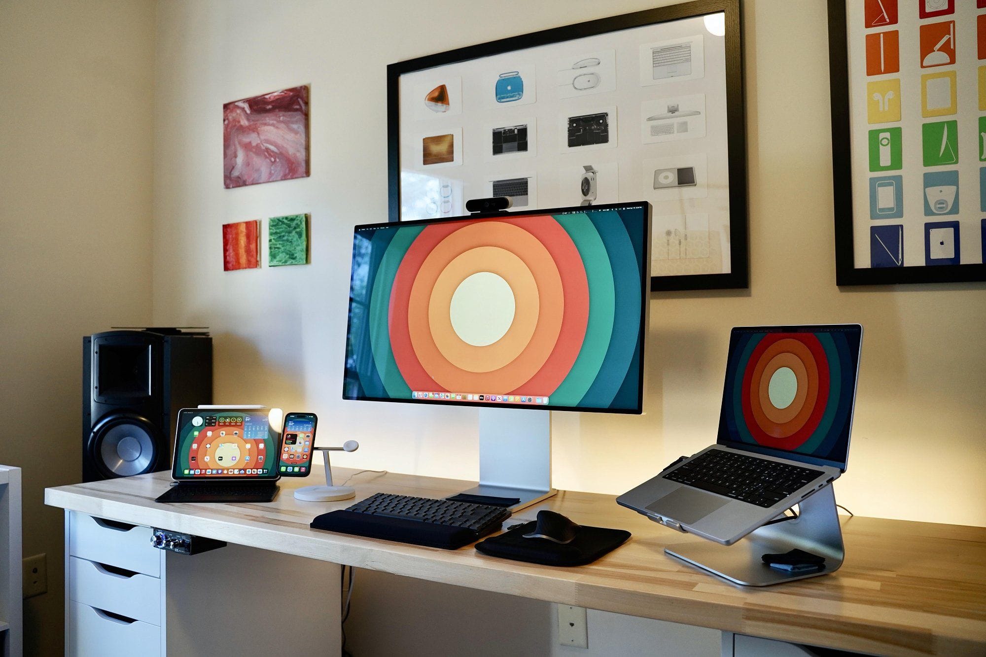 A contemporary Apple desk setup, accompanied by an iPad, iPhone on a charging stand, and a MacBook on a raised platform, flanked by abstract art and framed designs on the wall, on a wooden desk with a white drawer unit