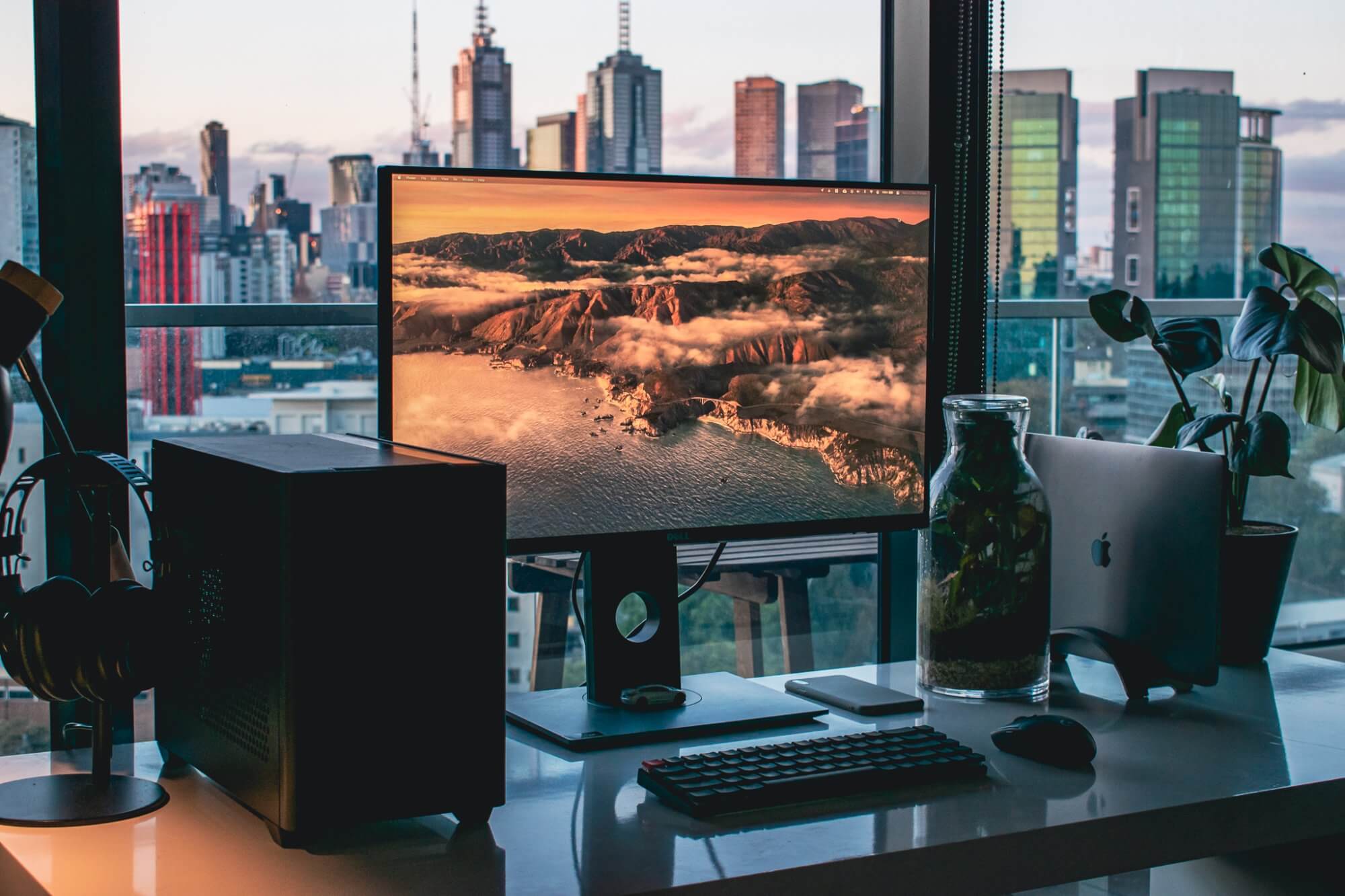 A minimalist desk setup with a sleek monitor, terrarium, and MacBook, overlooking an impressive city skyline through large windows