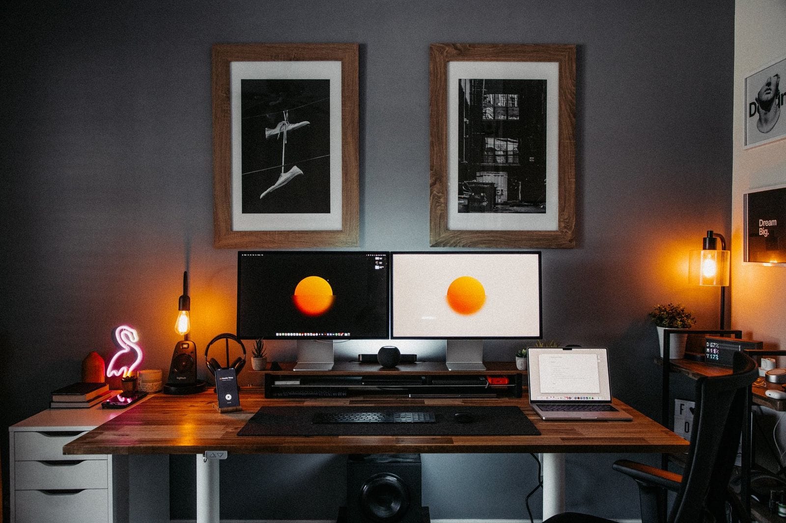 A home office setup with dual Apple Studio Display monitors on a wooden desk, featuring a neon flamingo lamp, a hanging Edison bulb, framed black and white photographs on the wall, and ambient lighting