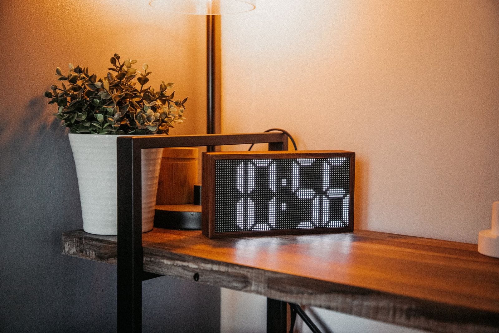 A wooden desk with a modern retro-style Tidbyt display showing digital art, flanked by a potted plant and a desk lamp, creating a cosy and tech-savvy workspace