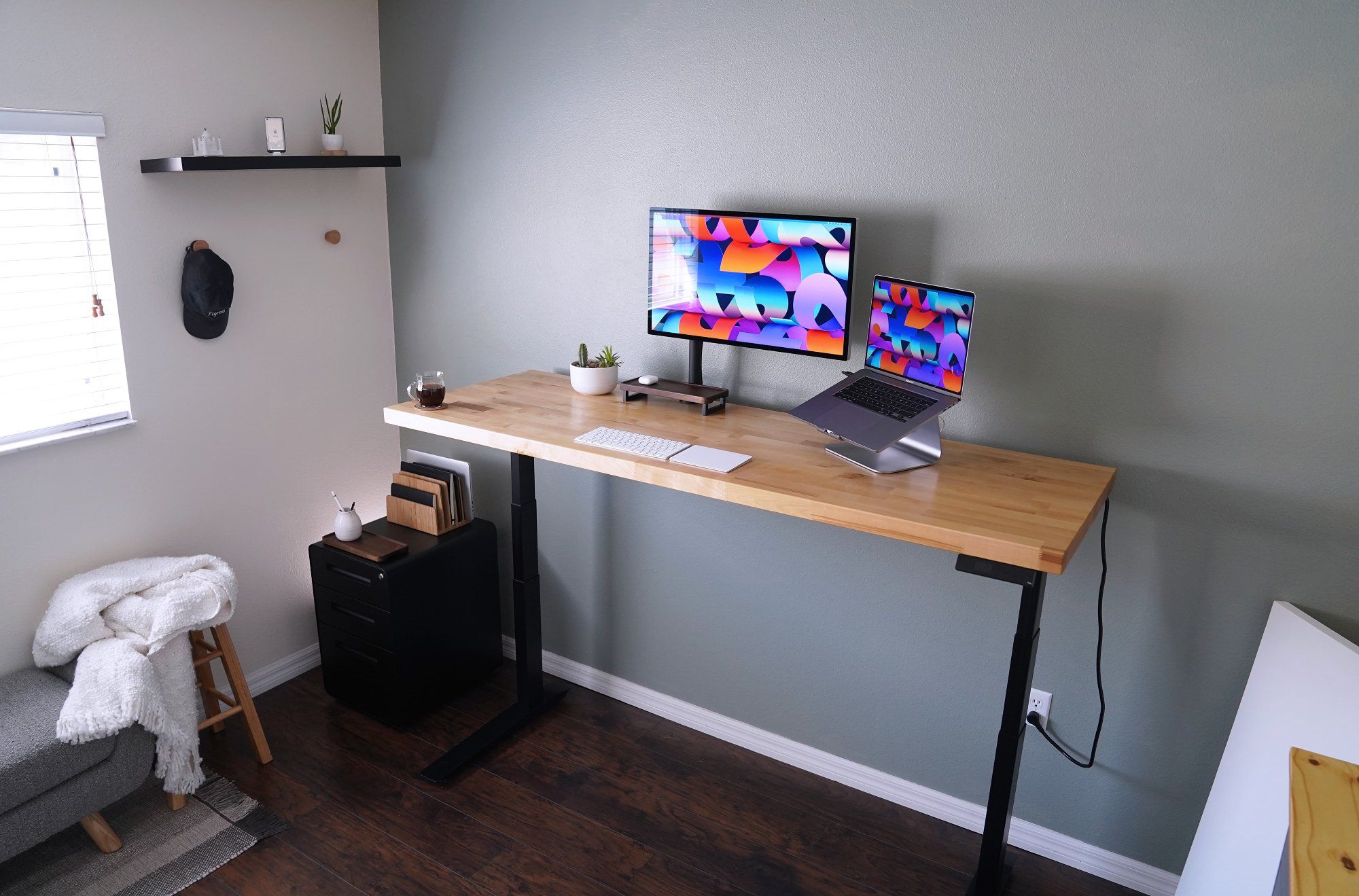 A minimalist workspace with a large wooden desk, single monitor, laptop on a stand, an armchair with a throw, and decorative floating shelf in a room with grey walls