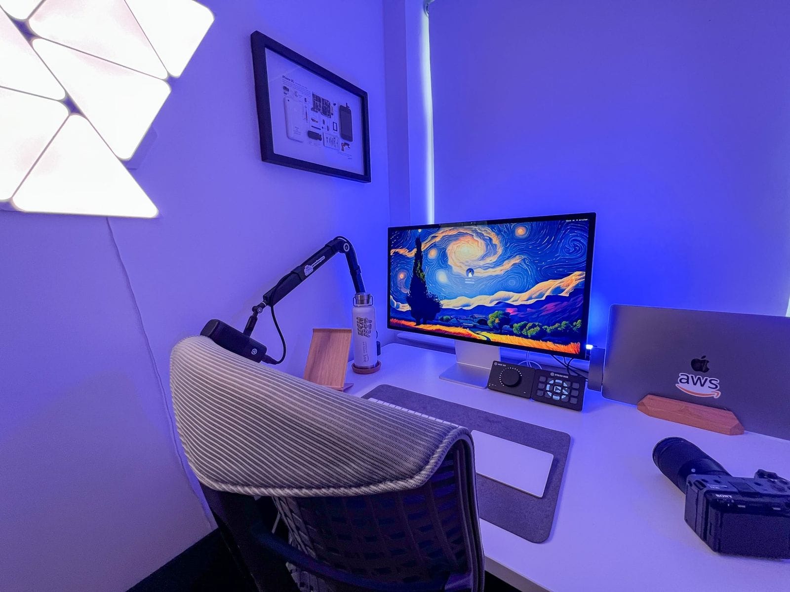 A modern home office setup illuminated by ambient blue lighting, featuring an Apple Studio Display with a vibrant screensaver, a MacBook with AWS branding, ergonomic chair, desk lamp, and a framed artwork on the wall, all arranged on a clean desk