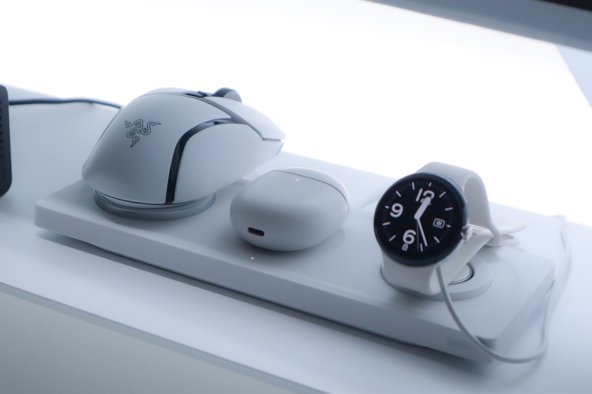 A high-precision gaming mouse, wireless earbuds in a charging case, and a smartwatch with a classic watch face displayed, all neatly placed on a white charging dock