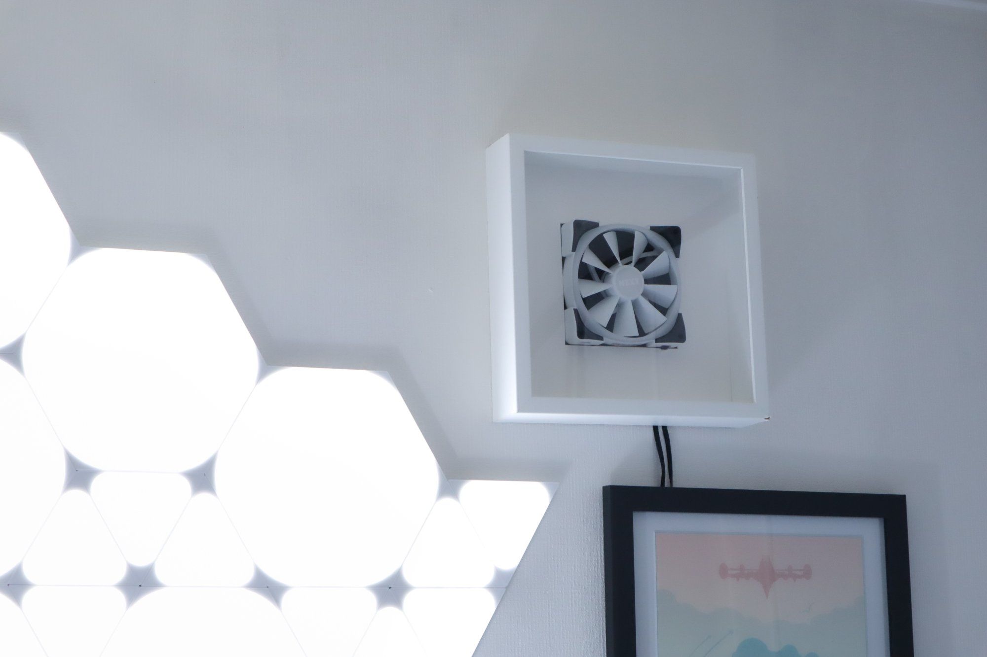 A minimalistic wall-mounted frame featuring a white PC fan, set against a modern backdrop of hexagonal light panels and a framed artwork