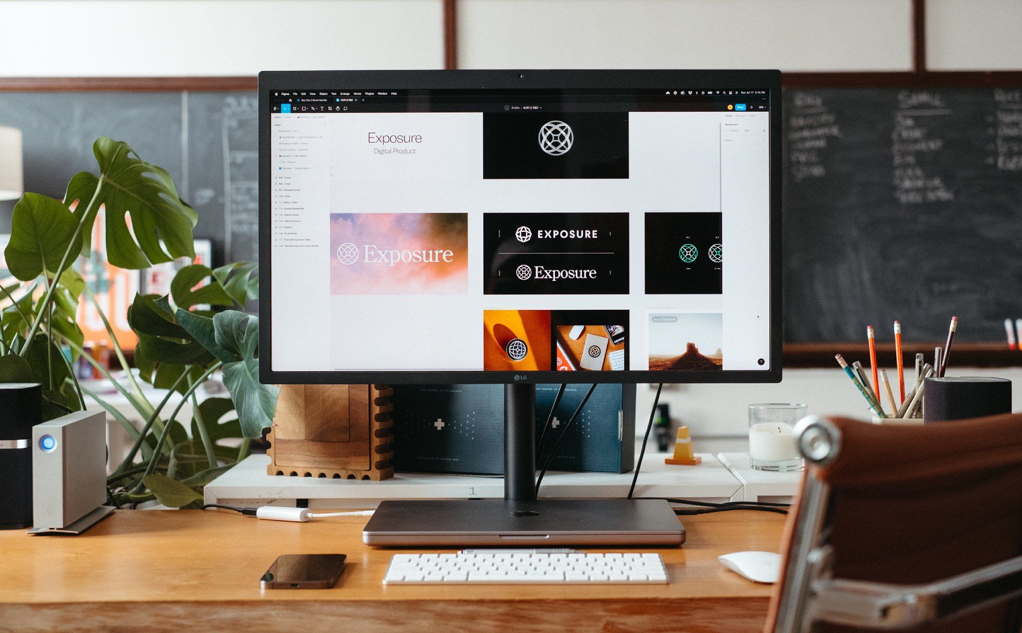 A minimal workspace with a wooden desk, large monitor, laptop, and classic leather chair in an old school building, with lush plants adding a touch of greenery