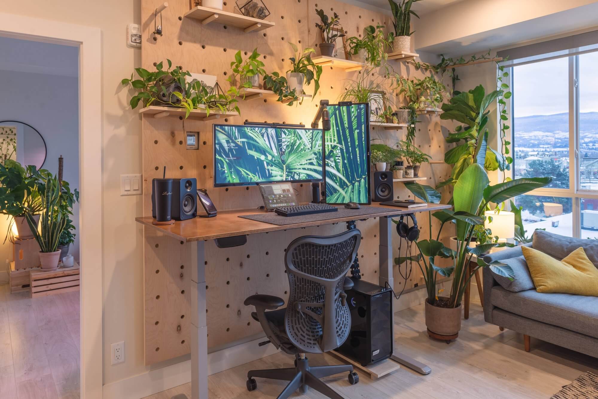 An ergonomic home office with a height-adjustable desk, dual monitor setup, speakers, and a variety of indoor plants on pegboards and shelves against a wooden backdrop