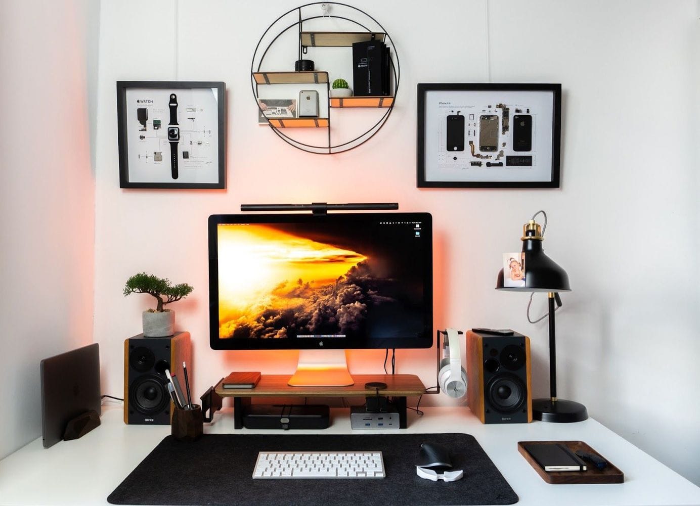 A modern workstation with an iMac displaying a vibrant cloud scene, flanked by speakers, a bonsai tree, and creative framed wall displays of disassembled Apple products, illuminated by a desk lamp and ambient lighting