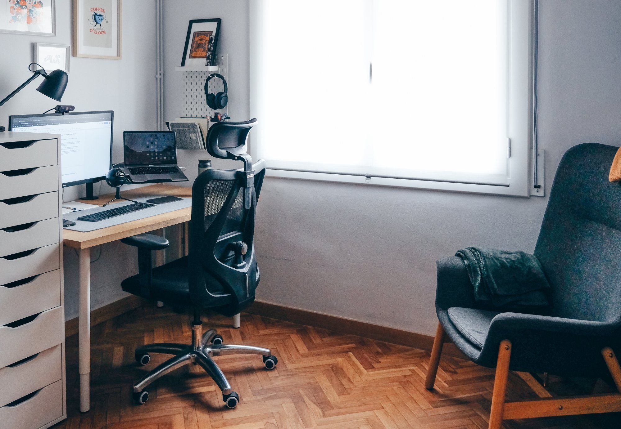 A minimalist home office with a light wood desk, ergonomic office chair, laptop with external monitor, pegboard with headphones, and a comfortable armchair with a throw blanket