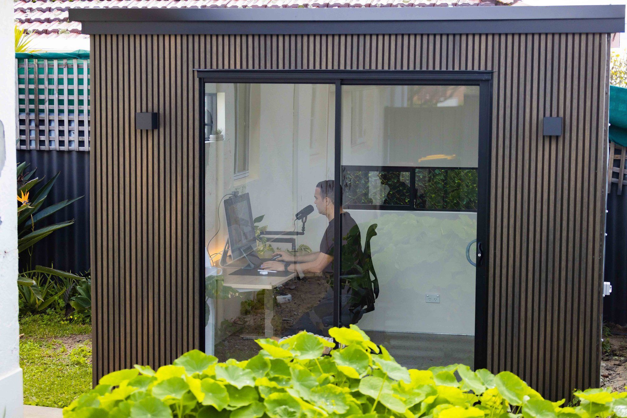A person sits at a desk inside a modern garden office pod with large glass doors, visible through the glass from the lush greenery outside. The office has a contemporary design with wooden slat walls and a flat roof