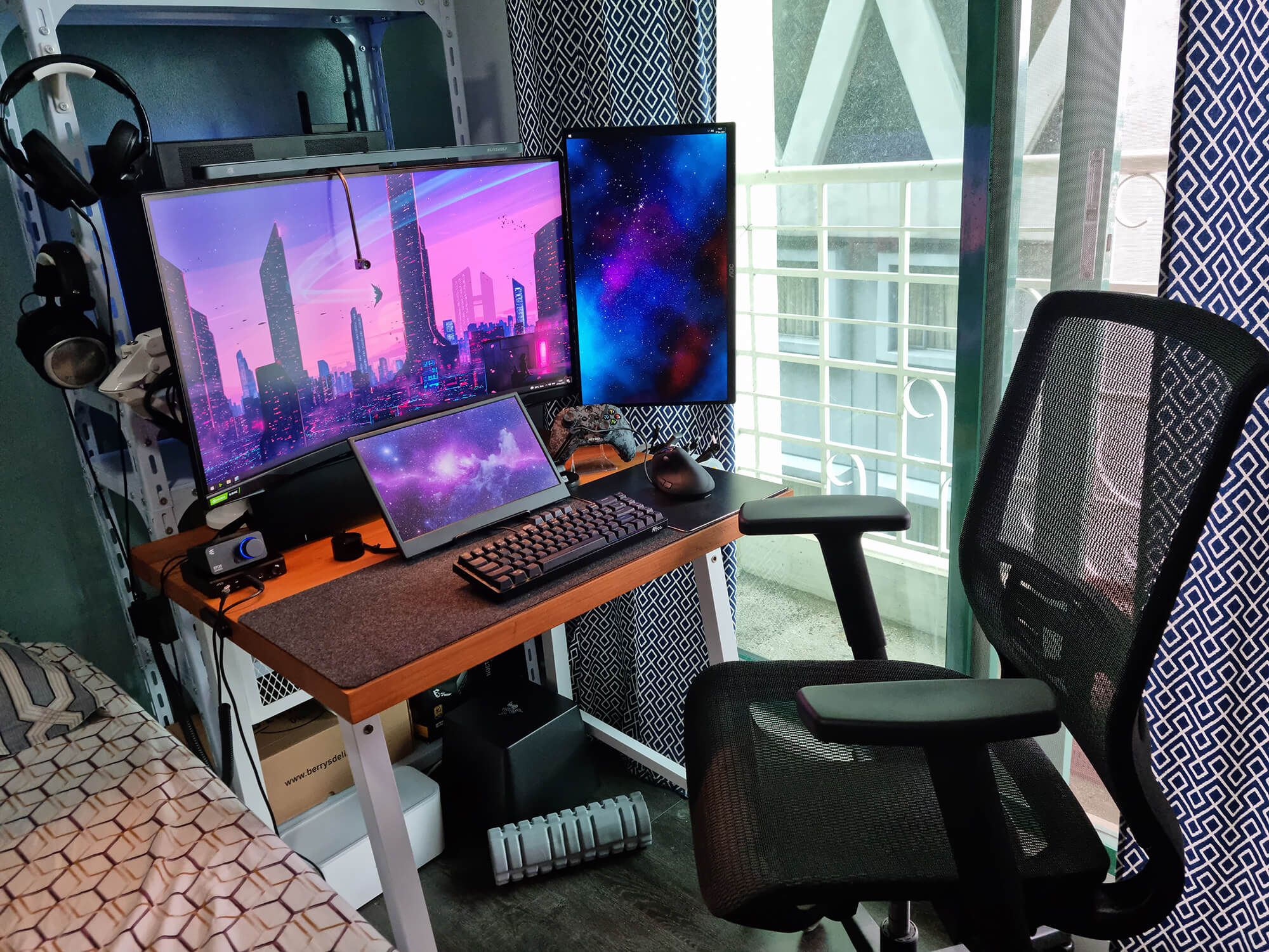 A home office setup with a large monitor flanked by a smaller vertical screen and a laptop on a wooden desk. The desk also has a keyboard, a mouse, a DAC (Digital-to-Analogue Converter), a headset hanging on the side, and a desk mat. There’s a mesh office chair in front of the desk and patterned curtains on the window to the right