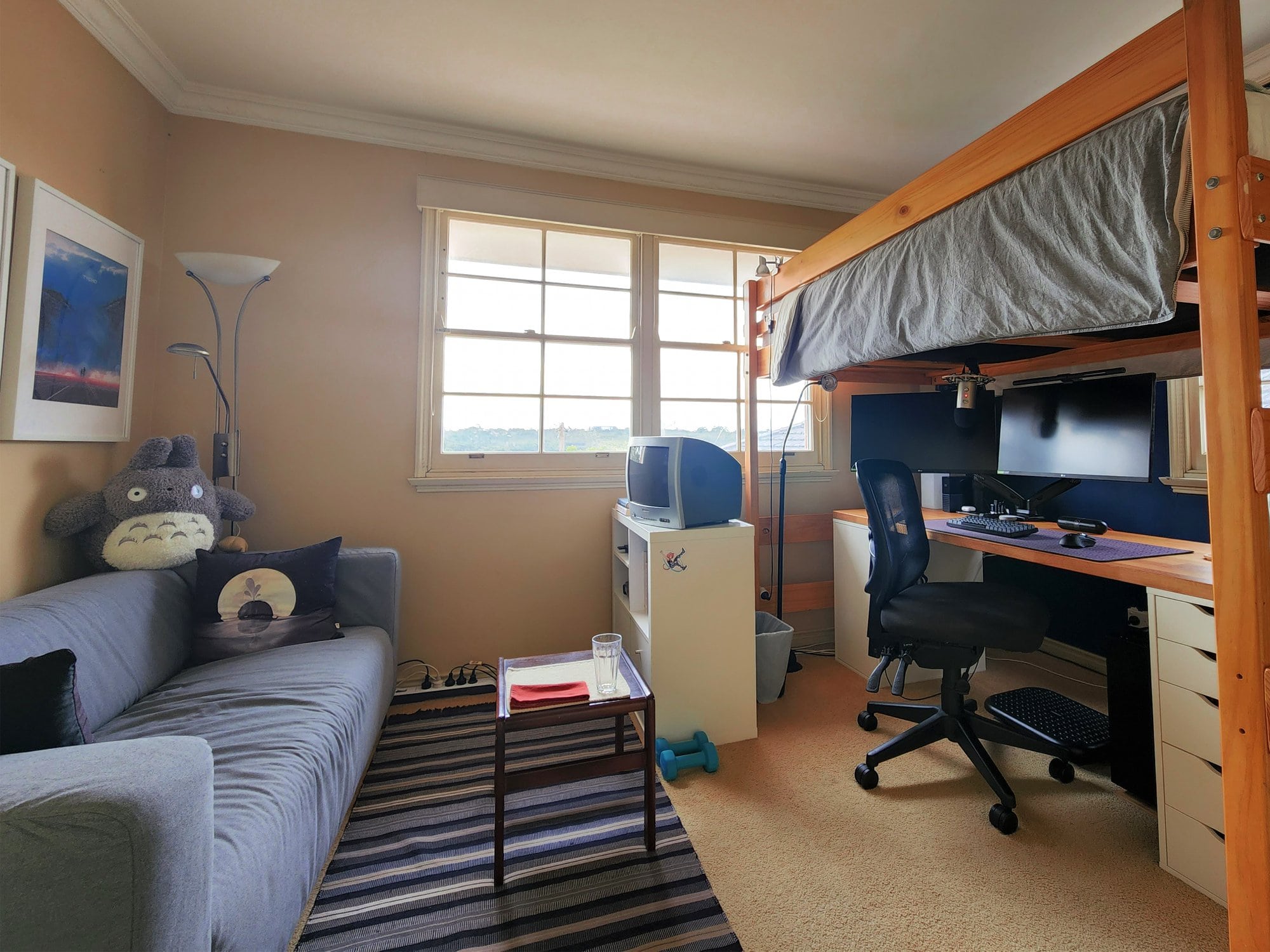 A cozy room with a loft bed above a workspace, which includes a desk with a computer setup and an ergonomic office chair. To the left, there’s a couch with a large plush toy and a small side table with a glass and a book. The room has a window, striped rug, and a standing lamp