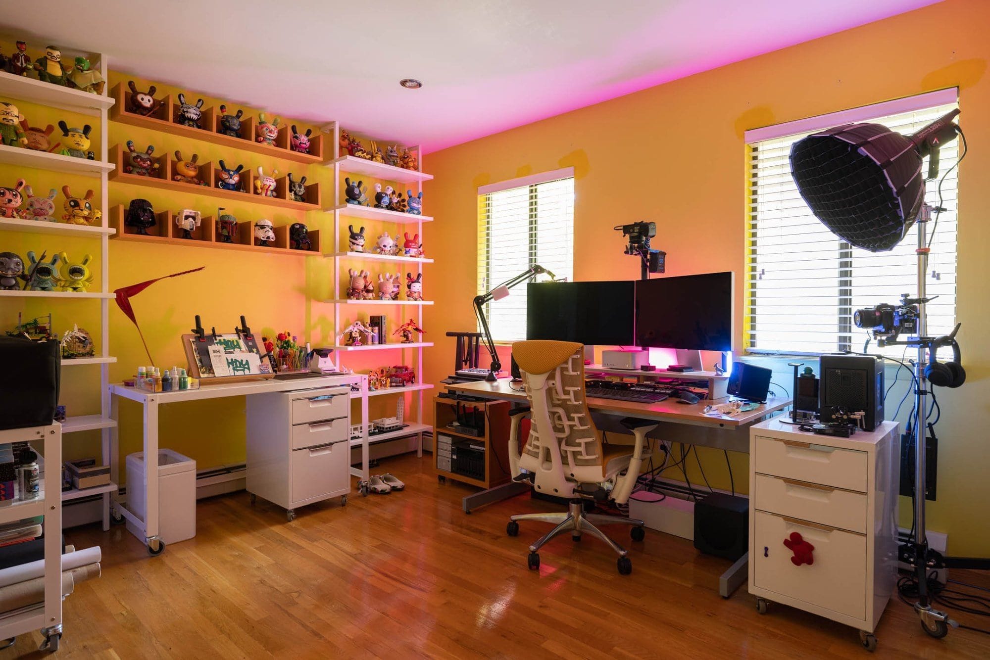 A vibrant creative workspace with wall-mounted shelves displaying colourful figurines, a desk with dual Apple monitors, a drawing tablet, a professional microphone and camera equipment, set against a wall with ambient pink lighting in a room with warm yellow tones