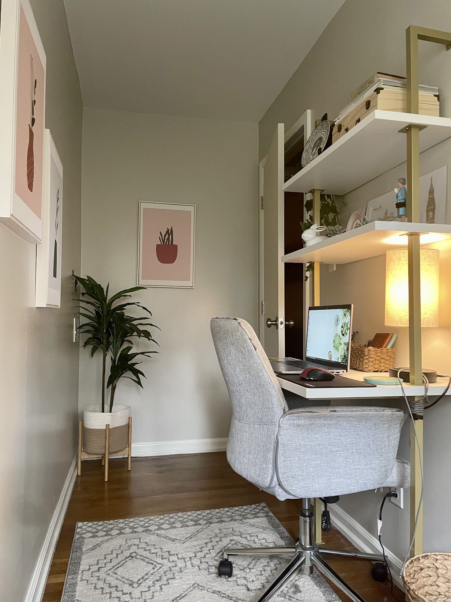 A tranquil home office featuring a grey upholstered swivel chair at a desk with a laptop, a lamp, and decorative items. Above, white open shelves display books, plants, and ornaments. On the left, artwork adorns the wall, and a potted plant sits in a white basket on the wooden floor, complementing a patterned grey rug