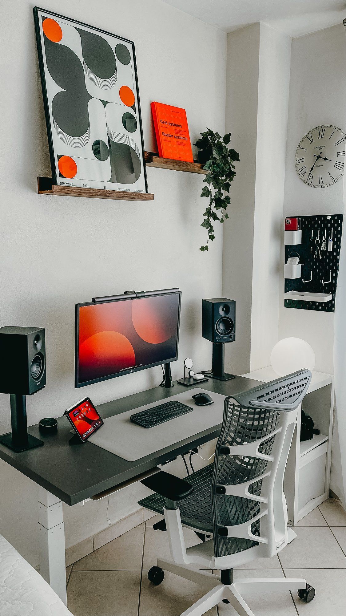 A minimalist bedroom desk setup with a grey adjustable desk, ergonomic mesh chair, dual speakers, and a black pegboard