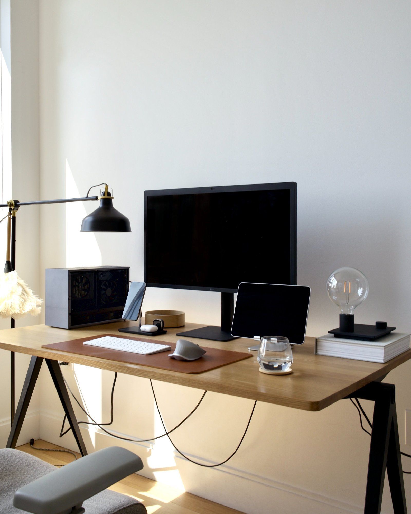 An Apple-like minimal desk setup with a sleek monitor, tablet, retro desk lamp, and a glass orb, all arranged on a wooden desk against a backdrop of minimalist decor