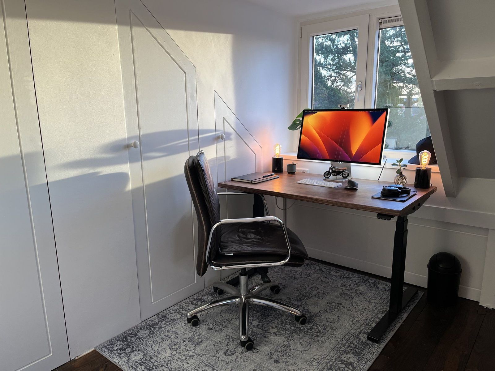 A simple attic home office setup with a height-adjustable wooden desk, leather office chair, large monitor, and decorative Edison bulb lamps, bathed in natural sunlight