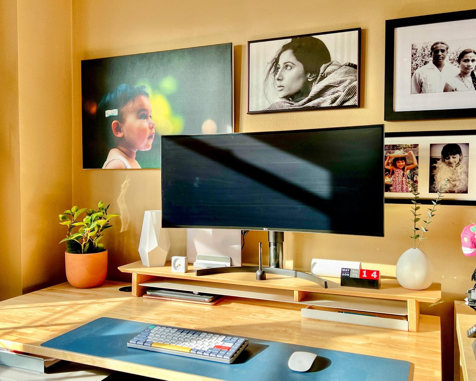 A home office desk bathed in golden sunlight, featuring a computer monitor, a plant, personal photos on the wall, and a desk calendar showing May 14th