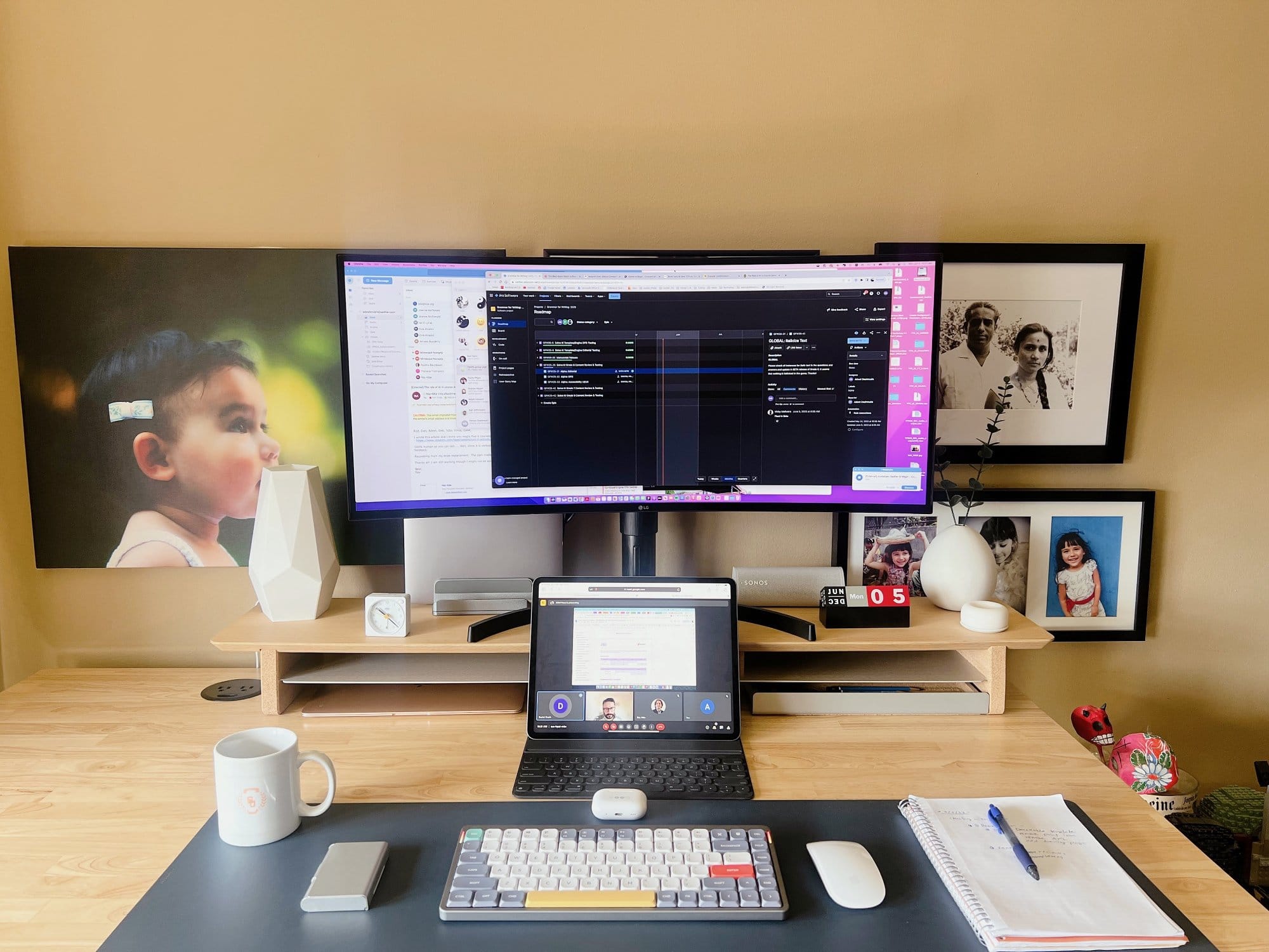A tidy home office space with a large monitor, a laptop, a mechanical keyboard, and family photographs on the wall, illuminated by soft natural light