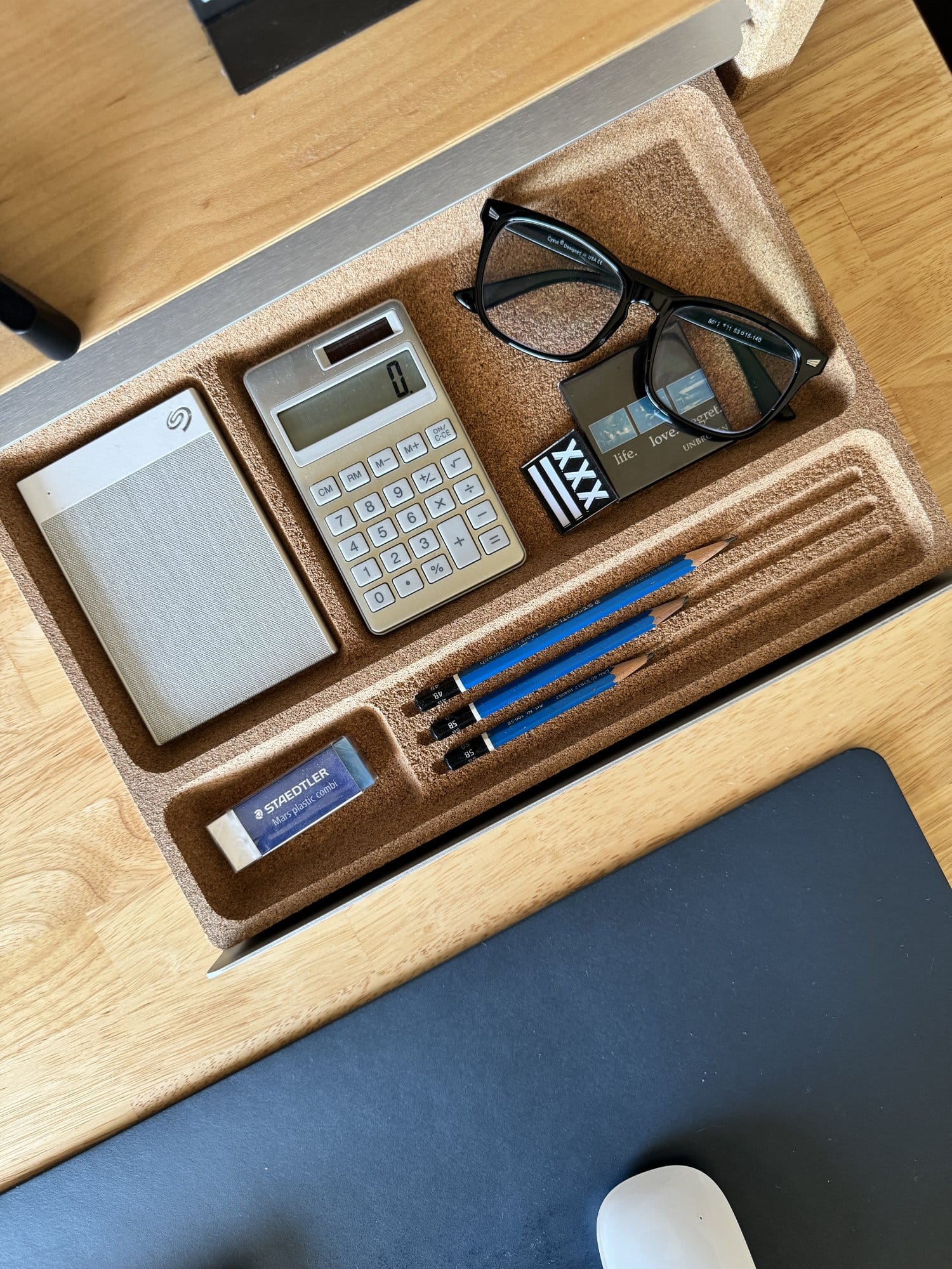 An overhead view of a desk organiser containing a calculator, spectacles, a small notebook, and blue pencils on a wooden desk next to a computer mouse