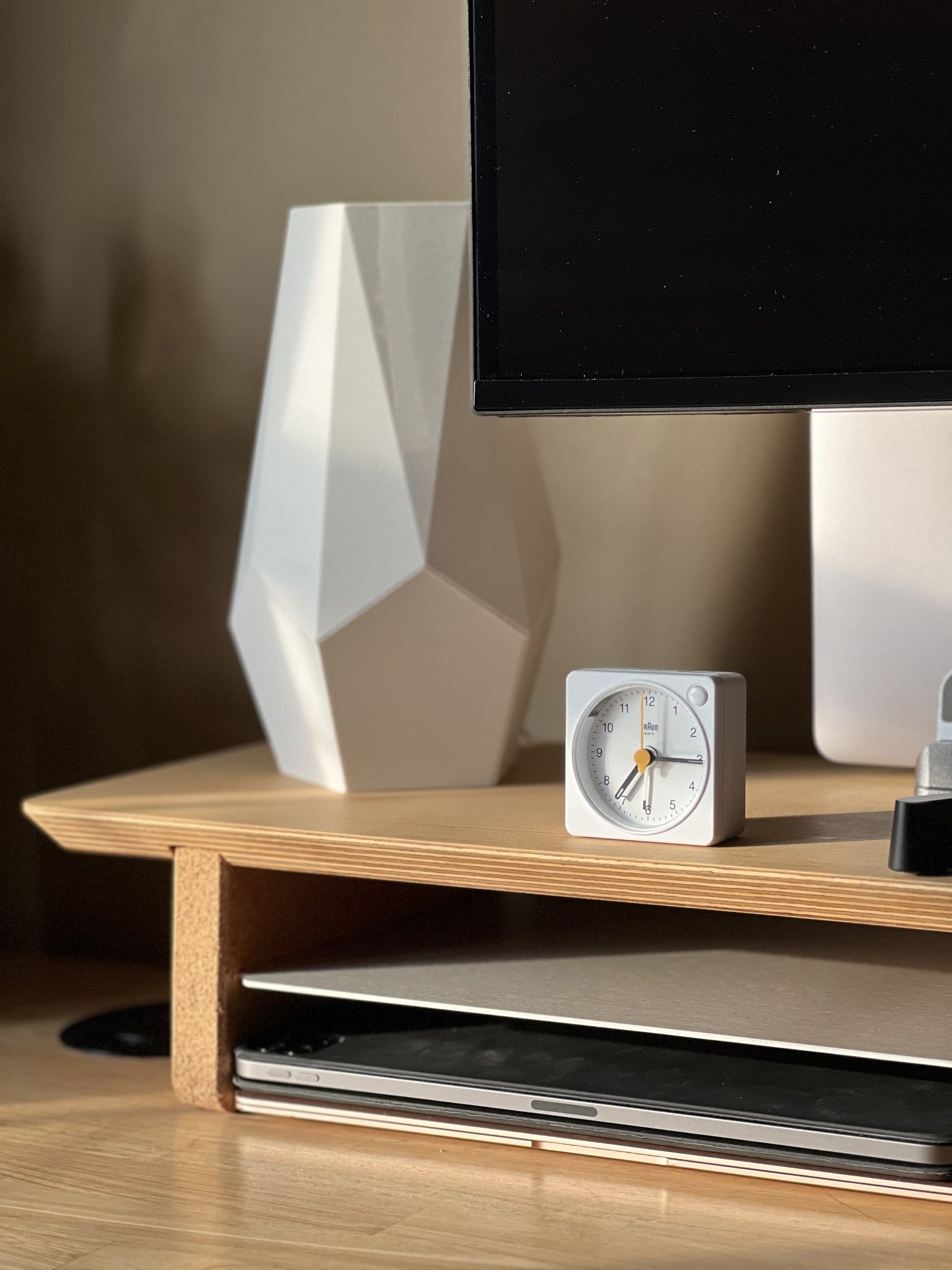 A close-up of a desk corner showing a modern geometric vase, an analogue clock, and a laptop in warm, natural sunlight