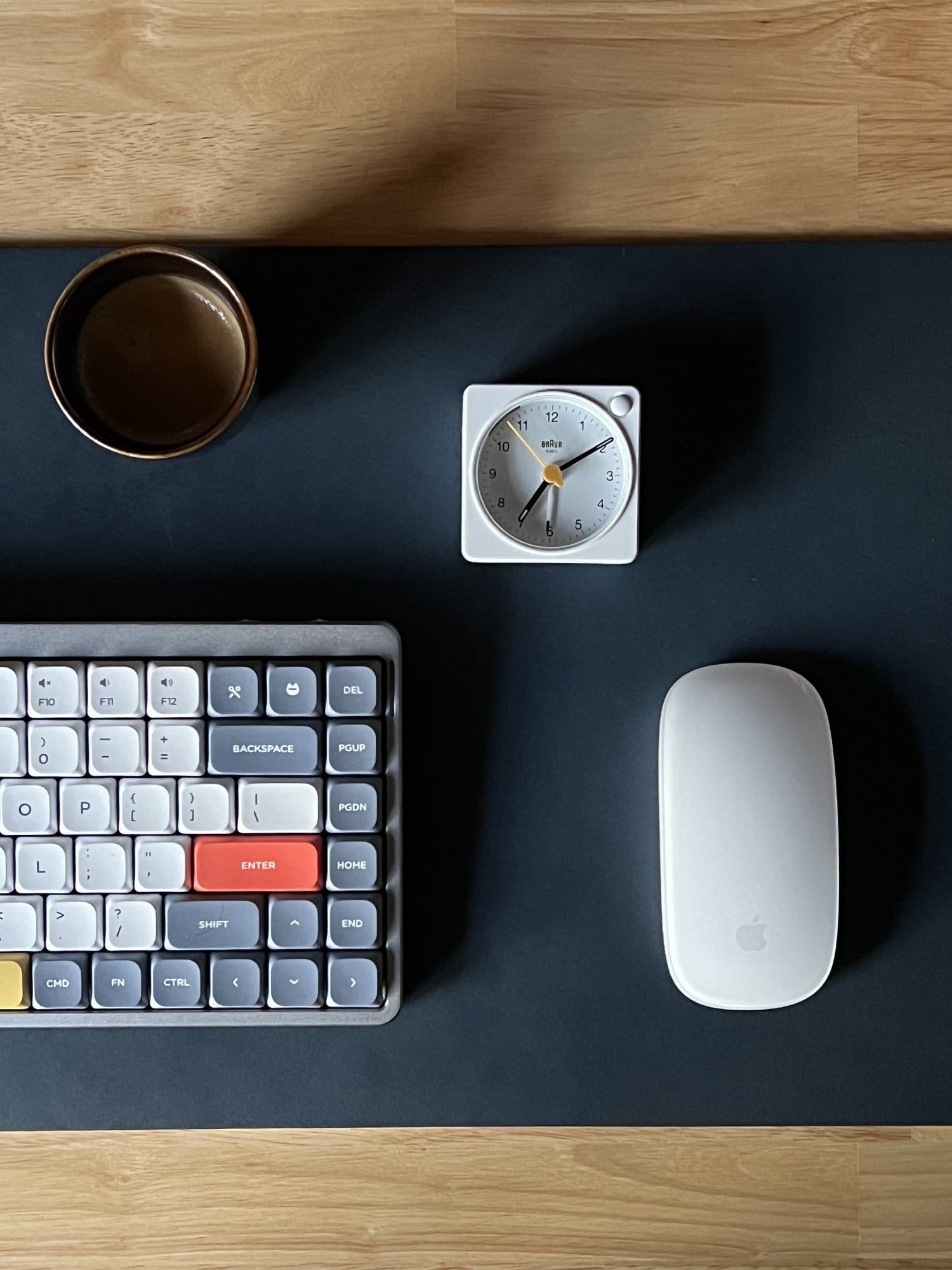  An overhead view of a desk with a classic mechanical keyboard, a white analog clock, a coffee cup, and an Apple mouse, all arranged on a dark mat