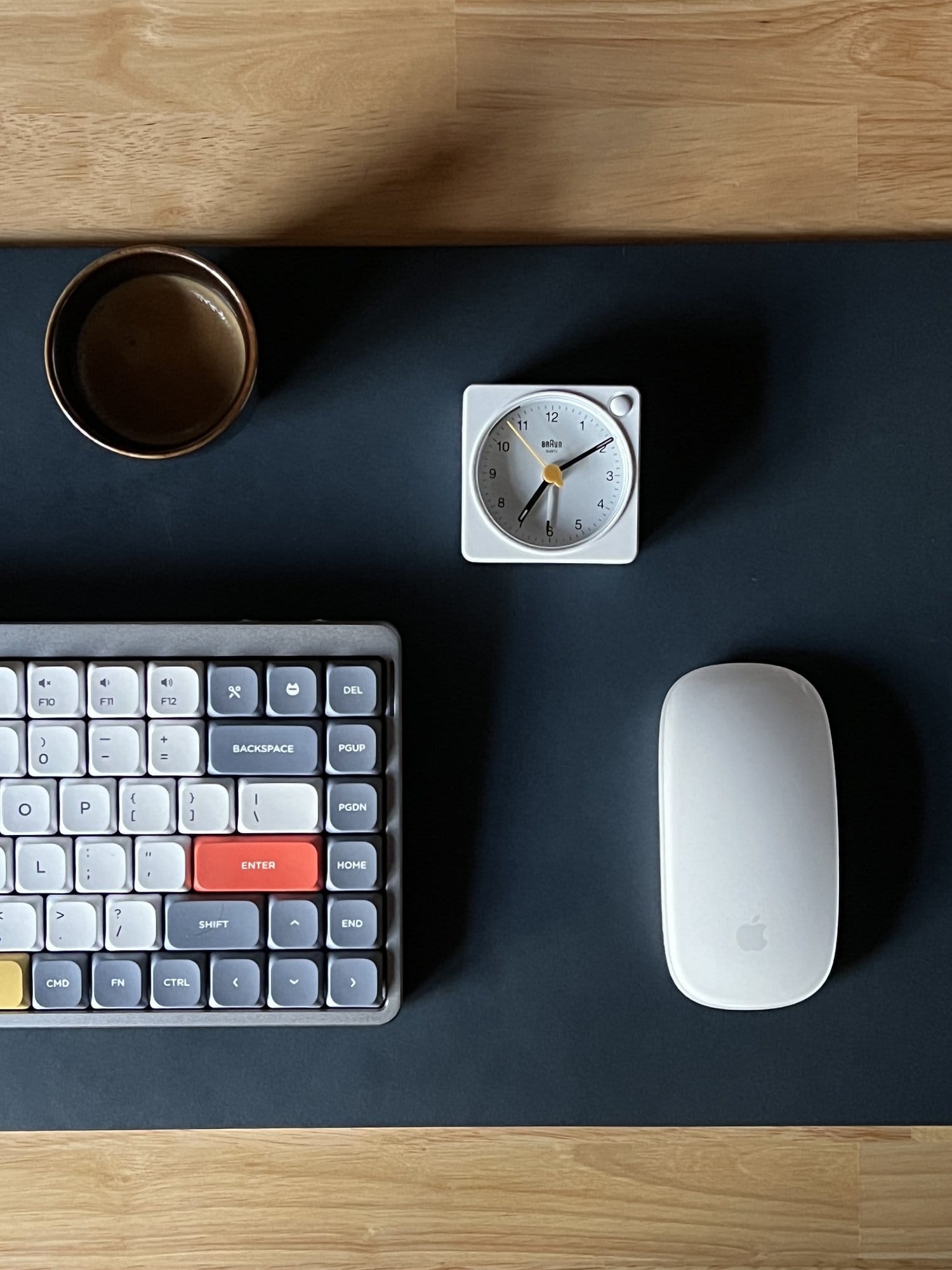 An overhead view of a desk with a classic mechanical keyboard, a white analog clock, a coffee cup, and an Apple mouse, all arranged on a dark mat