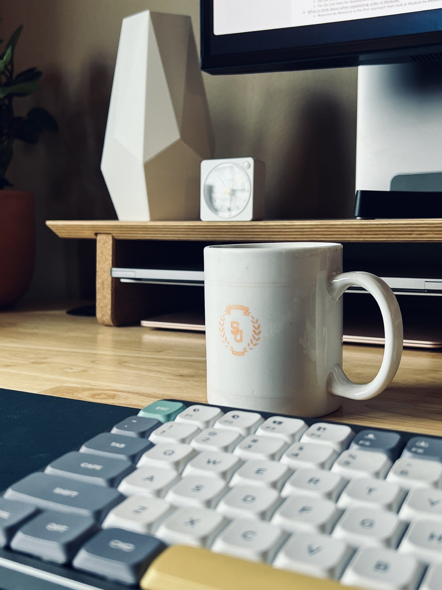 A close-up of a desk showing a part of a mechanical keyboard, a white mug with a logo, and a small clock, with a monitor and a geometric vase in the background