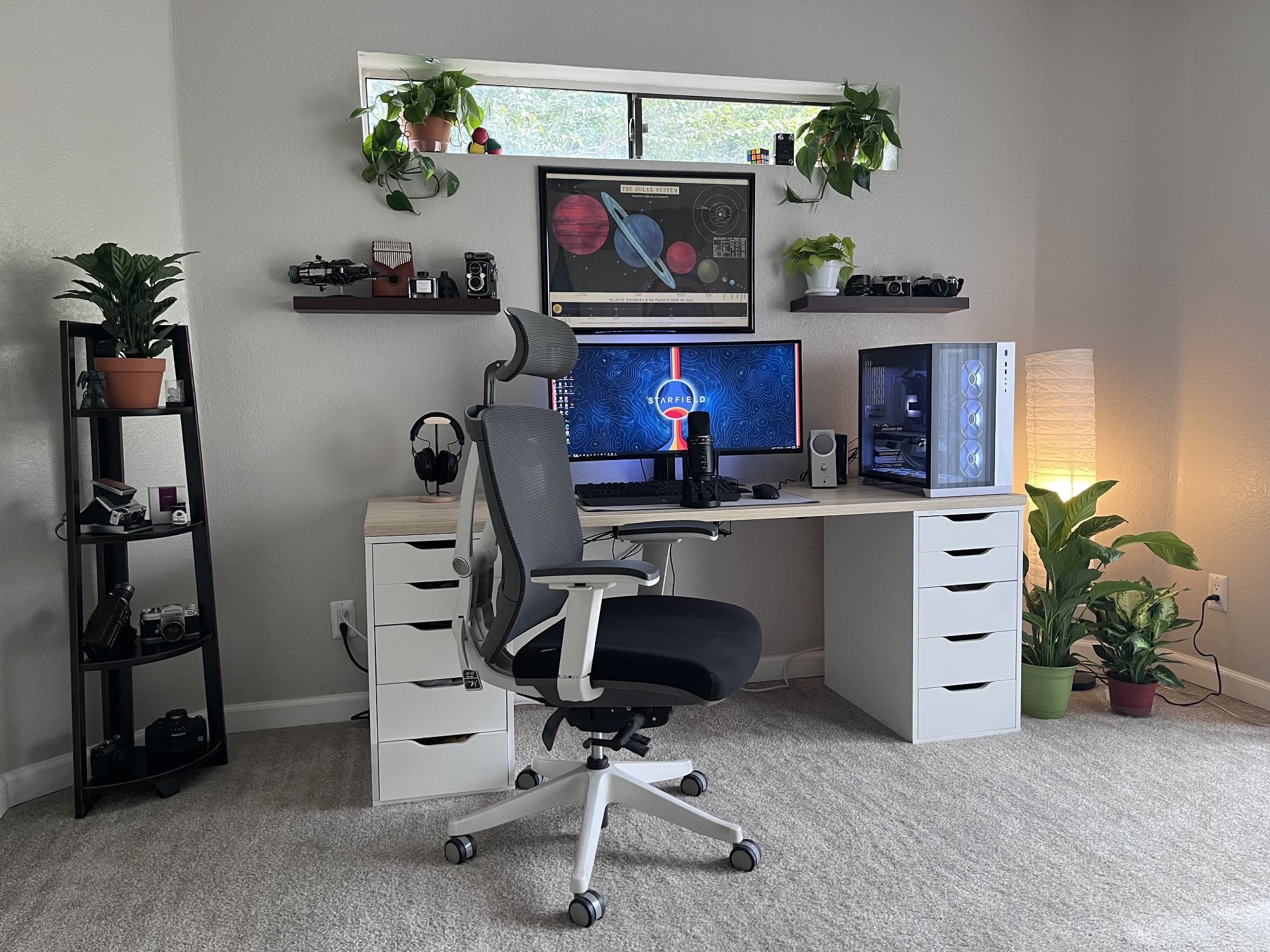 A home office setup featuring a white desk with a large curved monitor, a computer tower, houseplants, an ergonomic chair, and a shelf with photography equipment