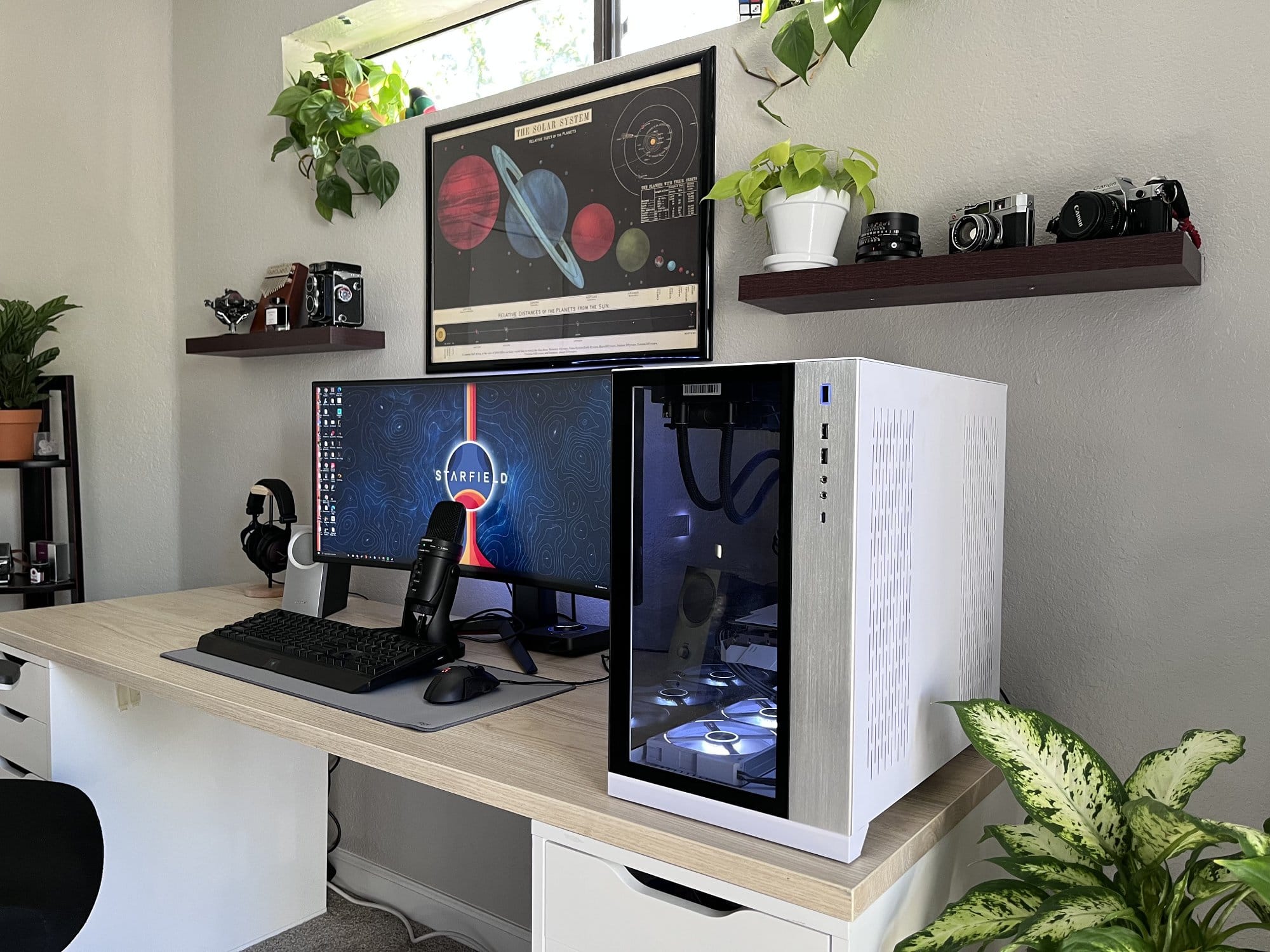 A modern desk setup with an ultrawide monitor displaying STARFIELD, a microphone, a gaming keyboard and mouse, a PC with a glass side panel, and decorative shelves with cameras and plants