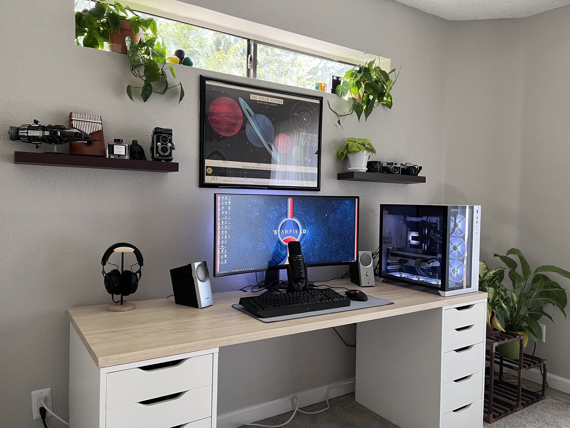 A clean and modern home office setup with a white desk, an ultrawide monitor, a gaming keyboard, a microphone, a speaker, a high-end PC, and shelves adorned with plants and vintage cameras