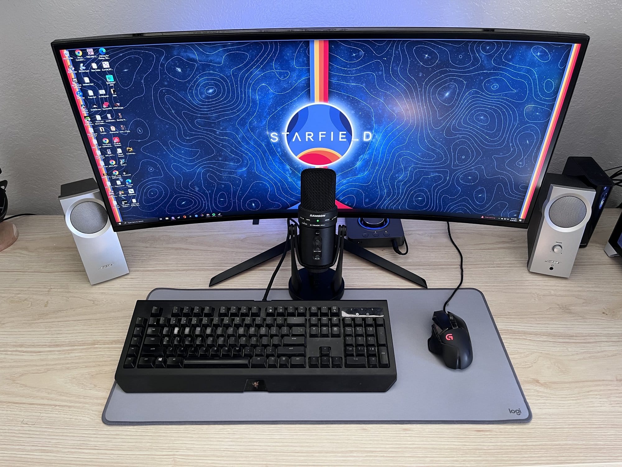 A curved monitor displaying a space-themed wallpaper with STARFIELD logo, flanked by stereo speakers, with a mechanical keyboard, gaming mouse, and professional microphone on a desk mat