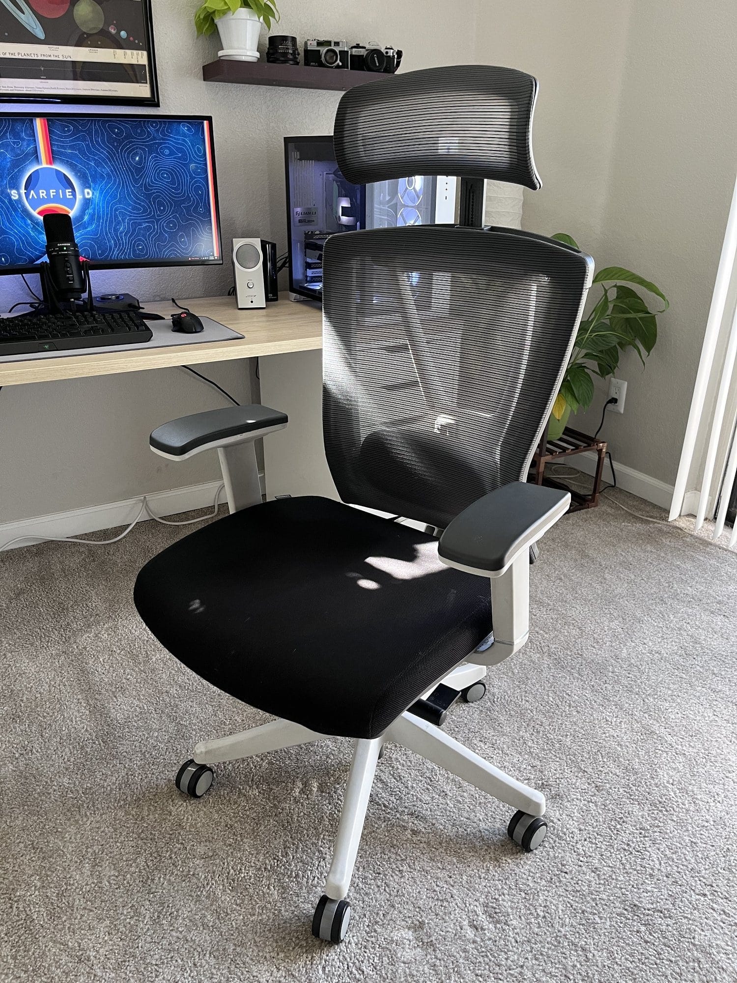 An ergonomic office chair with a mesh backrest and headrest, in front of a desk with computer setup and decorative shelves in a home office