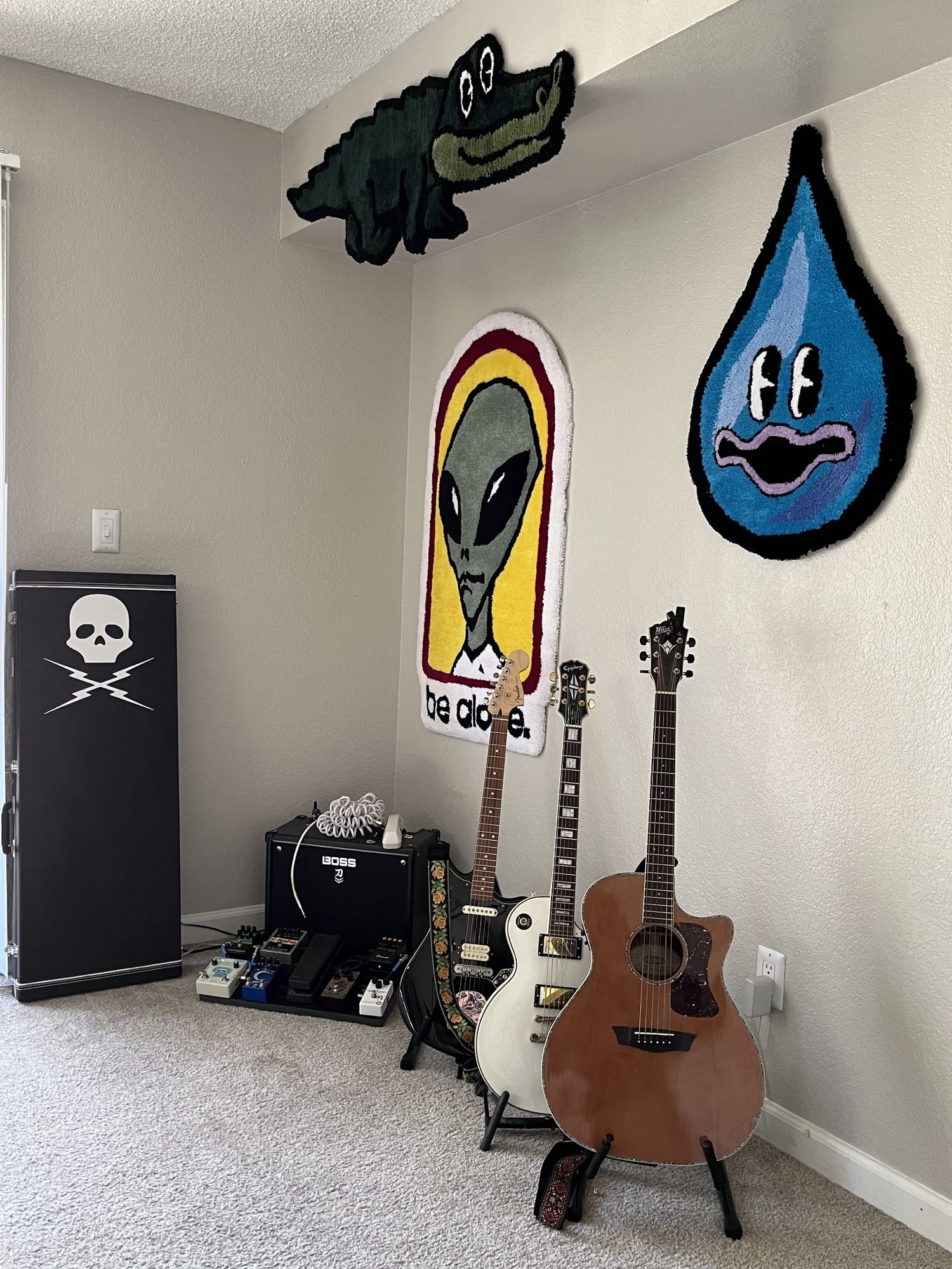 A music-themed corner with three guitars next to a BOSS amplifier, a pedalboard, and a storage case, under wall hanging rugs in the shapes of a crocodile, an alien, and a water drop