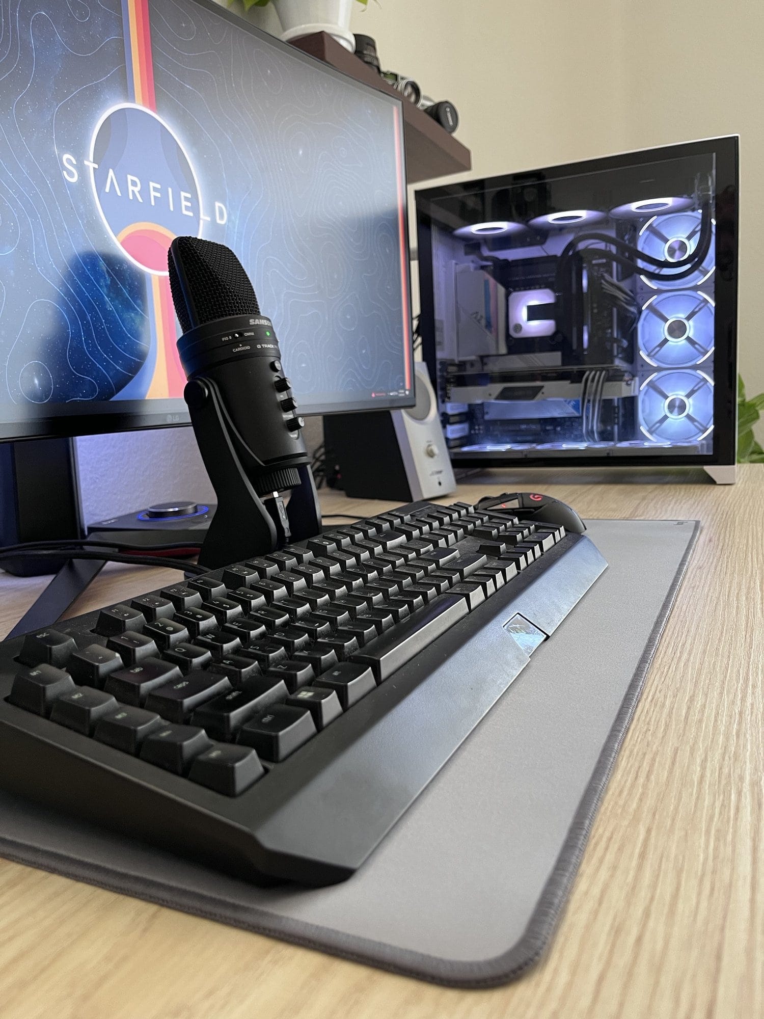  A close-up of a mechanical keyboard and a microphone on a desk, with a monitor displaying STARFIELD in the background and a PC with illuminated fans to the side