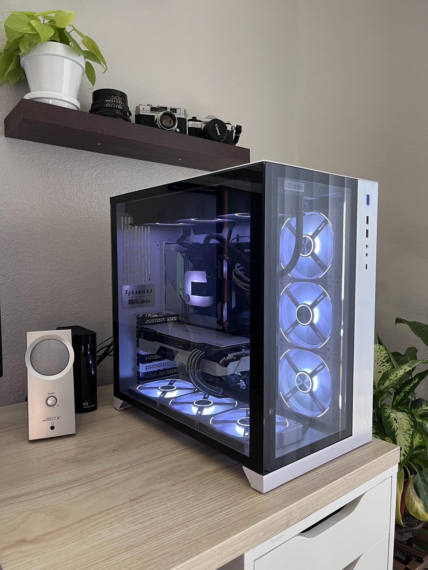 A high-end PC with a glass case and blue LED fans on a desk, flanked by a speaker and potted plant, with vintage cameras displayed on a shelf above