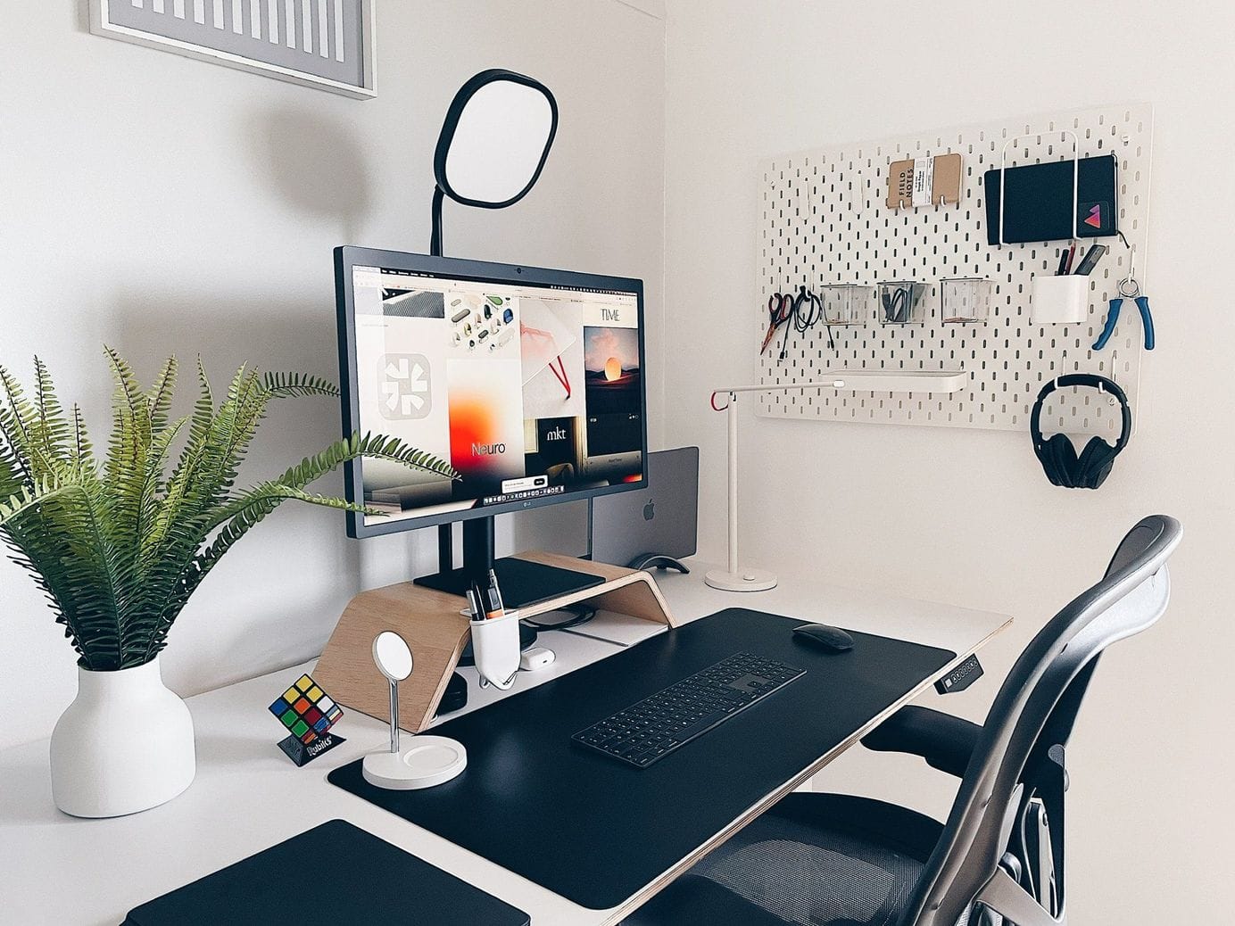 A minimalist desk setup with a monitor on a wooden stand, a potted fern, a pegboard with assorted office supplies, and an ergonomic chair, all neatly organised in a well-lit room