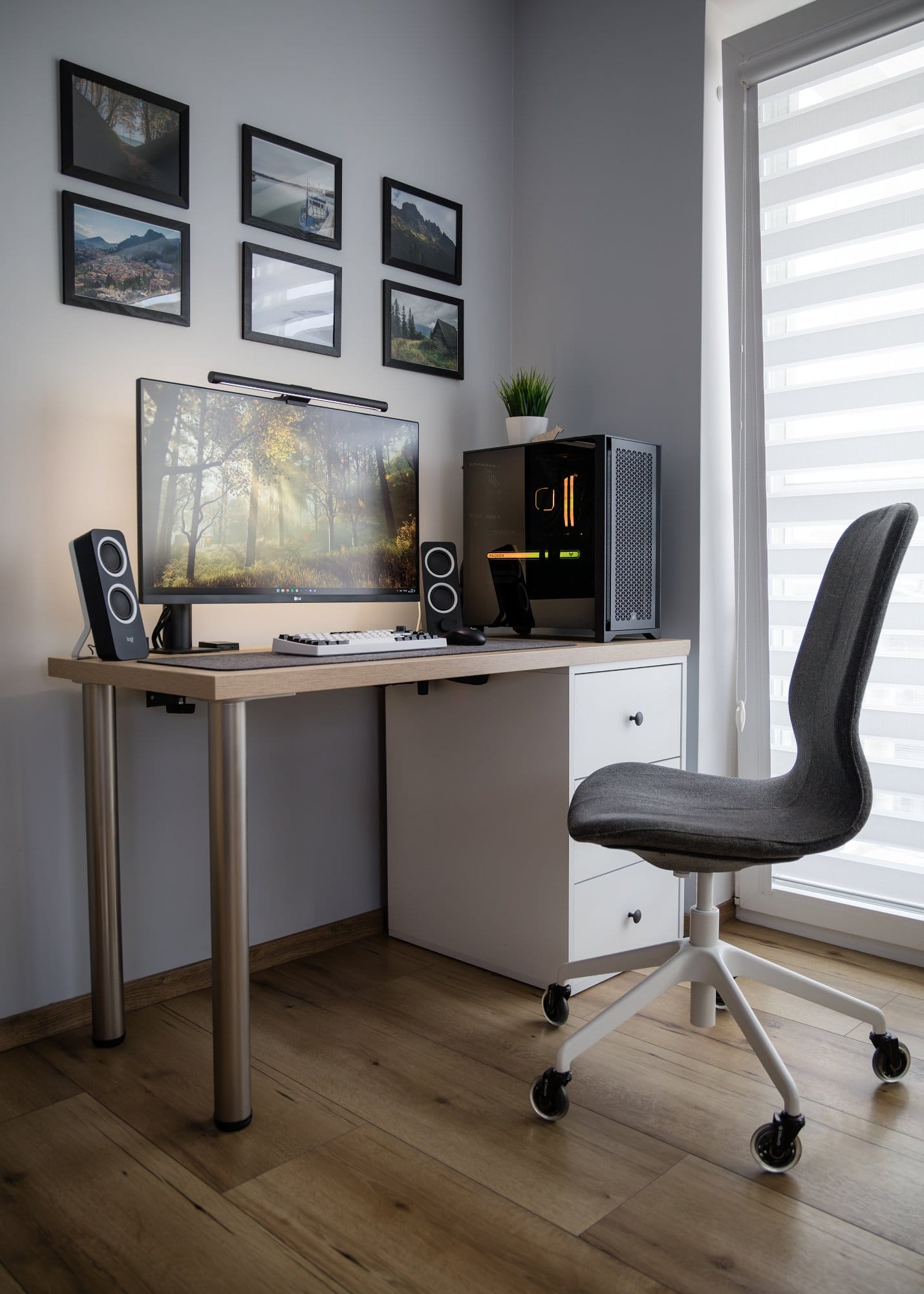 A contemporary home office setup with a corner desk, an ultra-wide monitor, speakers, a computer with illuminated elements, a grey IKEA swivel chair, and a backdrop of framed landscape photos